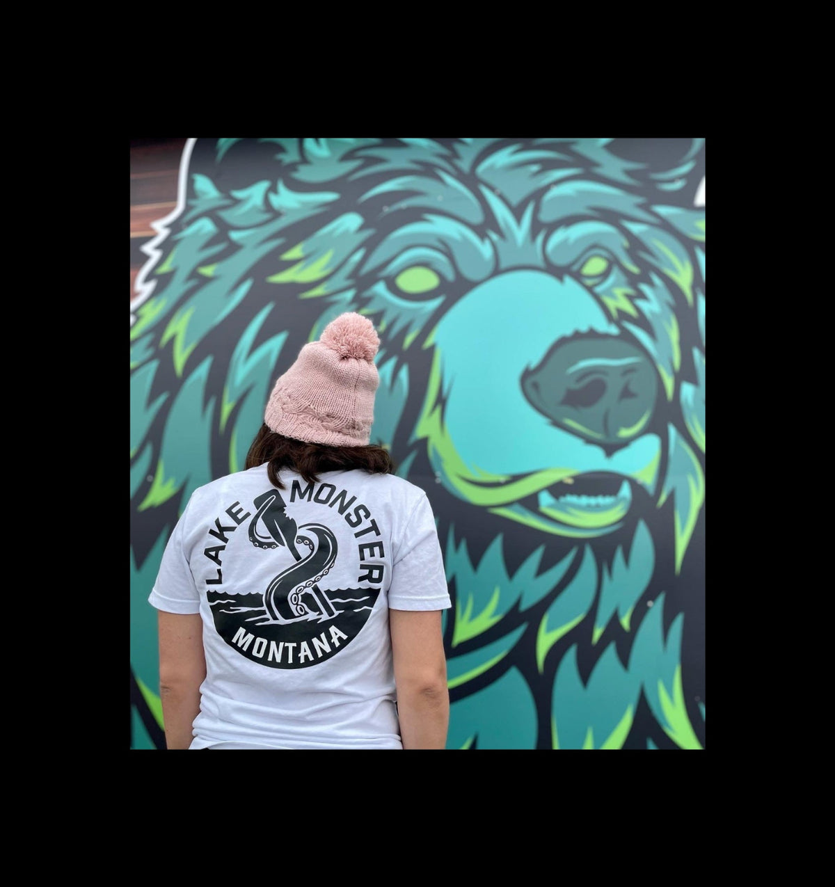 A person viewed from behind wearing a white graphic T-shirt and a pink pom-pom beanie, standing in front of a large mural of a blue and green bear face. The T-shirt has a black circular graphic reading 'LAKE MONSTER MONTANA' with a snake illustration.